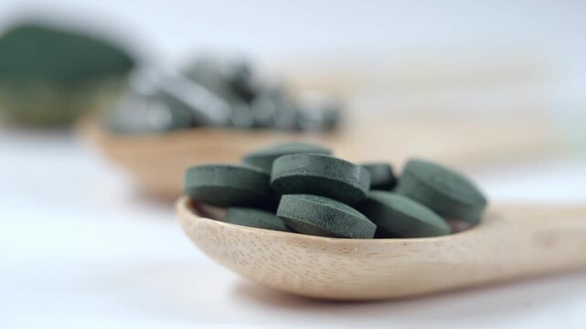 Macro footage of green spirulina and chlorella tablets on wooden spoon with blurred powder and capsules in background.