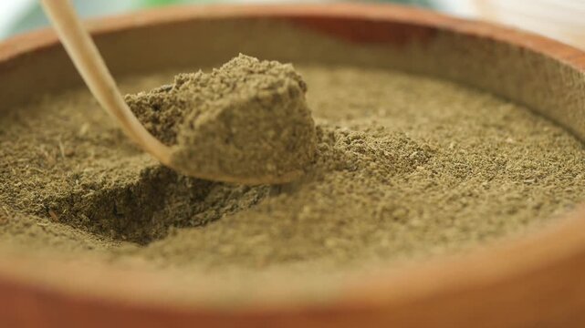 Close up of a wooden spoon taking scoop of fine cumin powder from a brown wooden bowl, showcasing aromatic Indian spice in motion.
