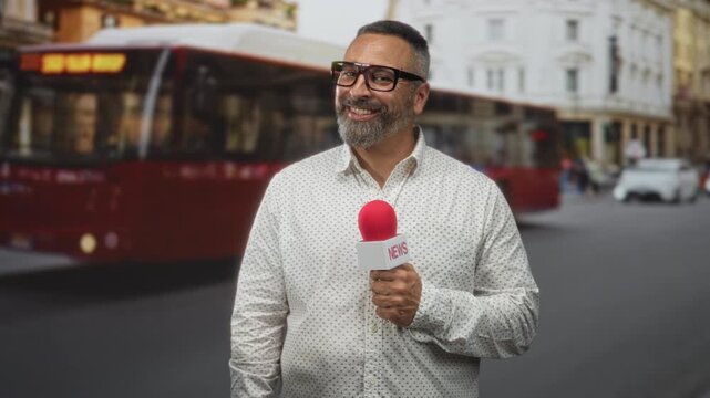Man reporter holding red microphone with news flag, hand gripping mic, wearing glasses and patterned shirt, grey beard visible, standing on a city street with bus and buildings behind; confidence.