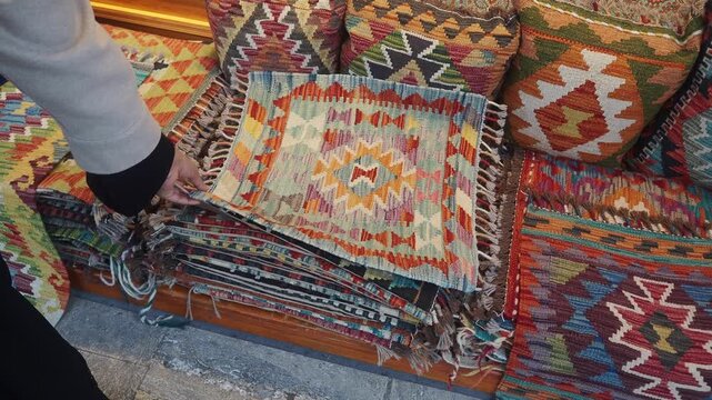 Person picking up colorful woven textile from stack of rugs and pillows.
