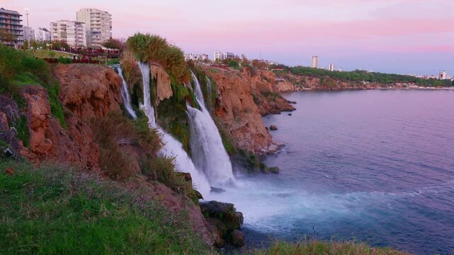 Coastal waterfall beside urban skyline at dusk, pastel pink sky, highrise buildings on cliff, crashing foamy pool, grassy promontory, seaside promenade and resort backdrop for sunset strolls