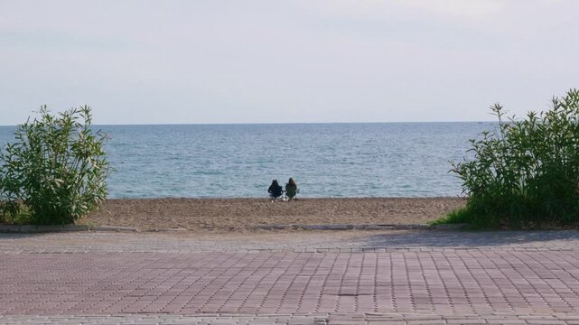 Serene scene on pebble beach. Peaceful depiction of lovers sitting beside calm sea waves. Silent moment captured with two individuals resting on pebble shoreline surrounded by nature
