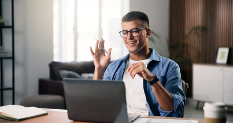 Obraz premium Education, laptop and video call with student man in home for online class, course or lesson. Computer, remote learning and wave with college or university pupil in apartment for virtual study