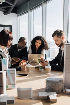 Diverse professionals collaborate around a table with laptops, tablets, and architectural models, discussing a project near large windows