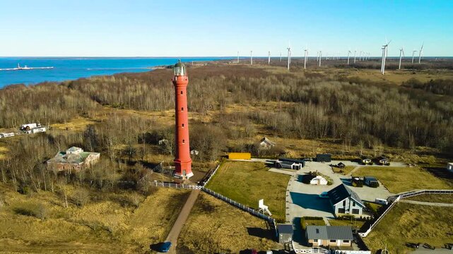 Sweeping aerial perspective of the historic red pakri lighthouse on a peninsula, with a modern wind farm generating renewable energy along the coastline under a clear blue sky in estonia