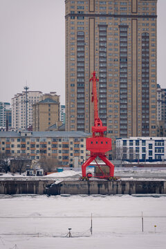 A snowy industrial port with cranes and stacked shipping containers set against urban buildings. Heihe, China