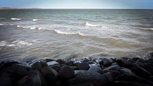 The rough sea drives waves against the grey stones of the coast under a cloudy sky, view from the west pier of the Baltic Sea near Puttgarden on 23769 Fehmarn, Germany