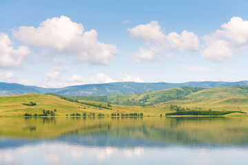 Green mountains and the blue sky with clouds are reflected in the lake. Beautiful summer landscape.