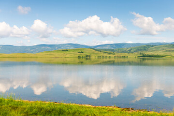 Green mountains and the blue sky with clouds are reflected in the lake. Beautiful summer landscape.