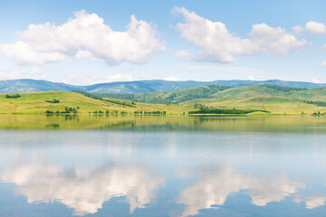 Green mountains and the blue sky with clouds are reflected in the lake. Beautiful summer landscape.
