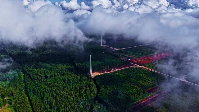 Drone footage captures tall wind turbines breaking through drifting mist above a mosaic of cultivated fields and rich forests