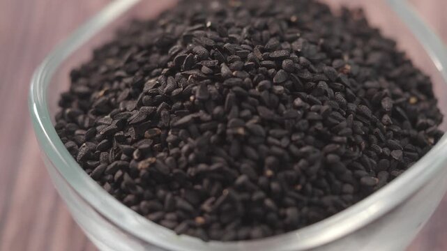 Close up of black cumin seeds in glass bowl on wooden background, macro shot of aromatic spice nigella sativa used in cooking and herbal medicine.