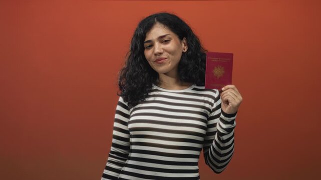 Young brunette woman holding passport and making ok sign to eye in studio; confidence travel identity.