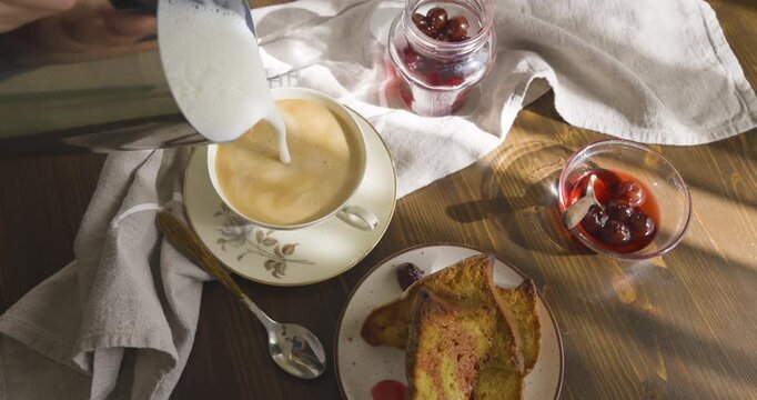 Pouring frothed milk into espresso cup to make cappuccino. Cozy morning breakfast with cake, cherry jam and warm sunlight shadows on rustic wooden table.