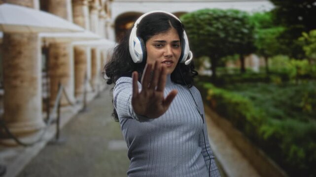 Woman wearing headset with hand covering eyes and palm raised in stop gesture on a stone colonnade street walkway; frustration.