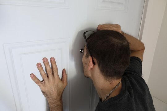 Close-up of a man pressing his face to a door peephole to look outside. Concept of curiosity, fear, and home security.