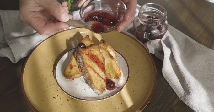 Anonymous person add cherry jam to pieces of cake on vintage ceramic plate with wooden spoon. Berry syrup in glass bowl, golden slices for cozy breakfast in natural sunlight.