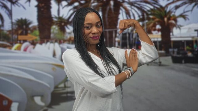 Woman flexes bicep beside stacked surfboards and palm trees on a sunny beach boardwalk wearing white blouse and bracelets; strength confidence.