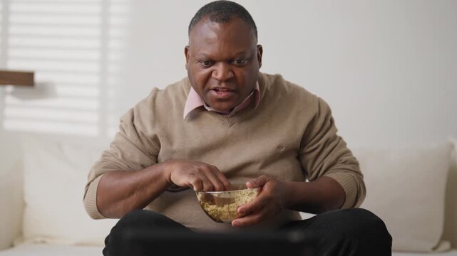 Sport fan watching TV and throwing popcorn when soccer team get goal, portrait. African american mature man relaxing in front of television set in living room in evening or in weekend, entertainment