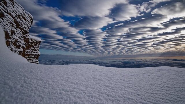 Snow covered cliff above cloud sea, dramatic altocumulus layers filling sky, textured snow foreground leading to distant ocean horizon, soft blue dawn light, rugged