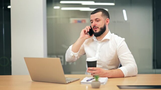 Smiling professional male sitting at office desk with laptop, talking on smartphone and holding coffee cup. Lawyer, IT developer or manager working in modern bright corporate workspace
