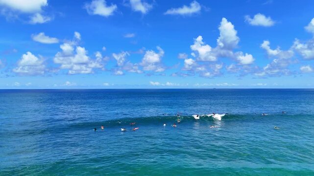 Aerial drone shot flying parallel to surfers riding waves near dramatic rocky cliffs in Bali, Indonesia. Dynamic action scene capturing the thrill of surfing in crystal clear ocean water