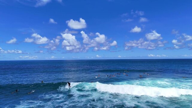 Aerial drone shot flying parallel to surfers riding waves near dramatic rocky cliffs in Bali, Indonesia. Dynamic action scene capturing the thrill of surfing in crystal clear ocean water