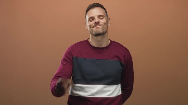 Young man in maroon and navy sweater tilting his head, smirking with hands behind his back against a brown studio wall; confidence ease.