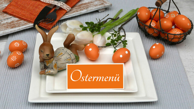 Tischdekoration zu Ostern. Ostertisch mit liegendem Hasen aus Holz, orangefarbenen Ostereiern mit wei&szlig;en Tulpen und einem Schild mit der Aufschrift 'Ostermen&uuml;'