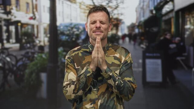 Man soldier in camouflage uniform with hands pressed together in prayer and closed eyes on a street; hope reflection calm.
