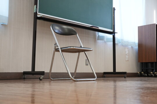 Empty classroom chair sits before a green chalkboard. Wooden floor reflects soft sunlight streaming through blinds. Simple metal frame supports dark plastic seat