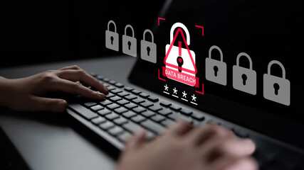 A person typing on a keyboard with a warning symbol on the screen signaling a data breach. This...