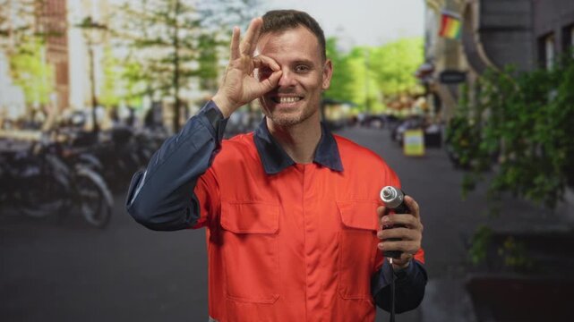 Man handyman in orange safety uniform holding drill and making ok sign over eye on a street with bicycles and buildings; pride skill.