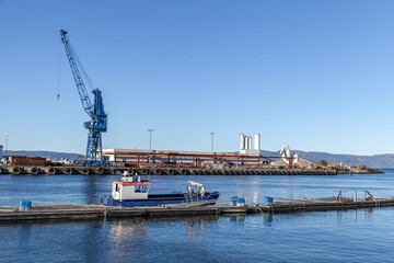 Obraz premium An industrial harbor scene featuring a large blue crane, a working boat moored at a wooden dock and coastal factory buildings along a calm waterfront beneath a clear blue sky. Trondheim, Norway
