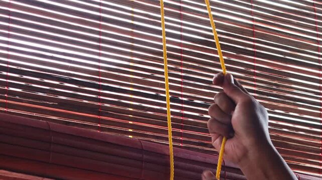 Close up of a man&rsquo;s hands pulling a rope to adjust bamboo blinds, allowing sunlight and fresh air to enter, improving ventilation and natural lighting in a home.