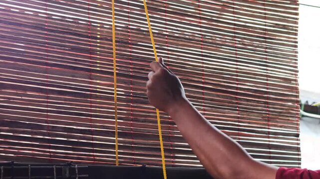 Close up of a man&rsquo;s hands pulling a rope to adjust bamboo blinds, allowing sunlight and fresh air to enter, improving ventilation and natural lighting in a home.