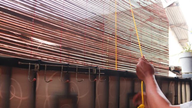 Close up of a man&rsquo;s hands pulling a rope to adjust bamboo blinds, allowing sunlight and fresh air to enter, improving ventilation and natural lighting in a home.