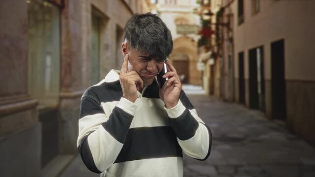 Man plugs left ear with finger while holding smartphone to his right ear on a cobblestone street; frustration communication problem.