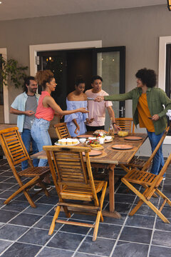 Diverse friends standing and leaning around wooden dining table on tiled patio, passing plates