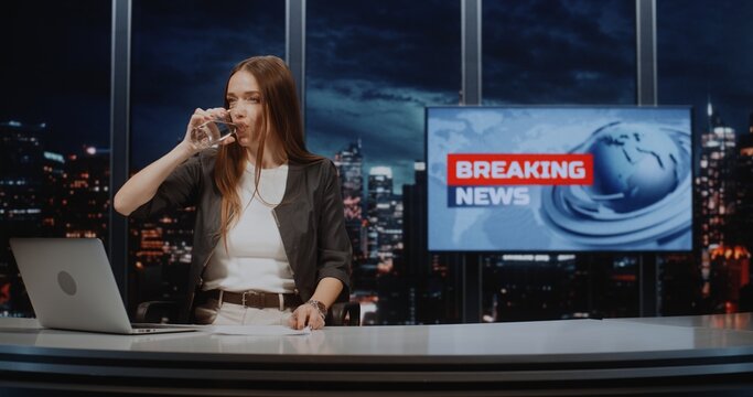 Female Anchor Sits at Desk Reviewing Papers and Drinking Water. Lighting Adjusted In Background. Concept Of Live Broadcast Preparation, Journalism, Professional Portrait In Modern High Tech TV Studio.