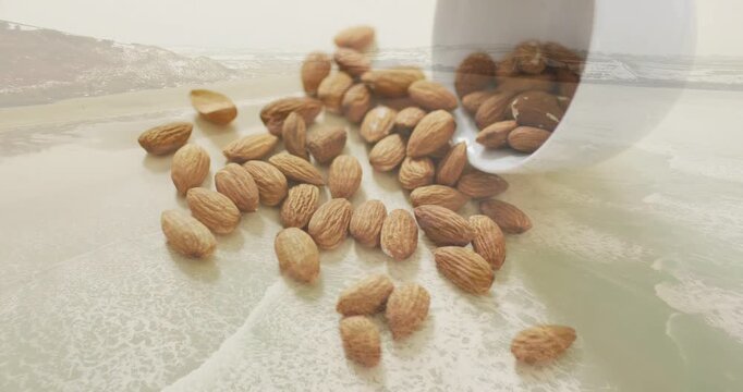 Cup tilting and pouring almonds onto pale surface, pile forming under coastal overlay for food ads
