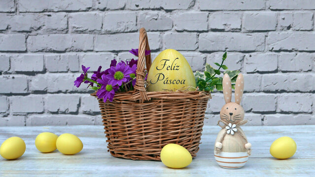 Uma cesta de vime com margaridas roxas e um ovo de P&aacute;scoa amarelo com a inscri&ccedil;&atilde;o portuguesa "Feliz Pascua" em frente a uma parede de tijolos brancos.
