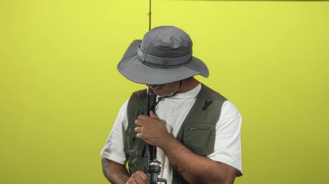 Man holding fishing rod with hands, tilting head and looking at reel while wearing sunhat and fishing vest in studio; serenity.