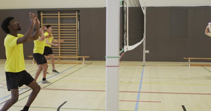 Diverse male team in yellow shirts drilling at school gym by net after lobbed volleyball passing