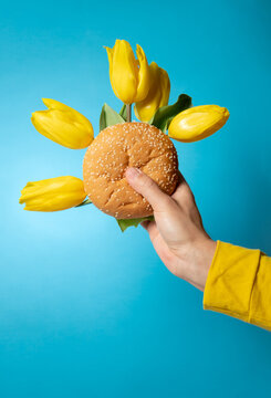 Hand holding burger with yellow tulips arranged on blue background