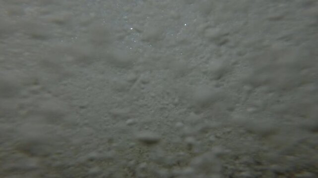 Sea waves quickly approaches the camera and covers it, wave moves pebbles on seabed, below surface the water mixes forming many air bubbles, Underwater split level,  Close-up, Slow motion