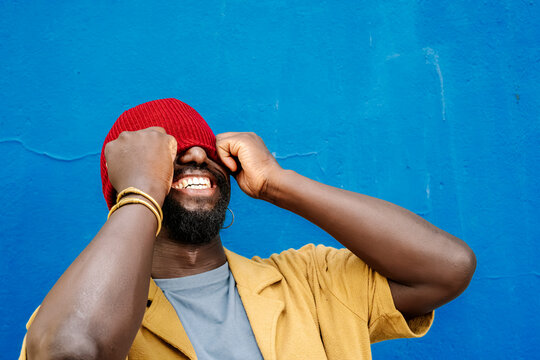 Smiling man with beanie laughing against blue wall in urban setting