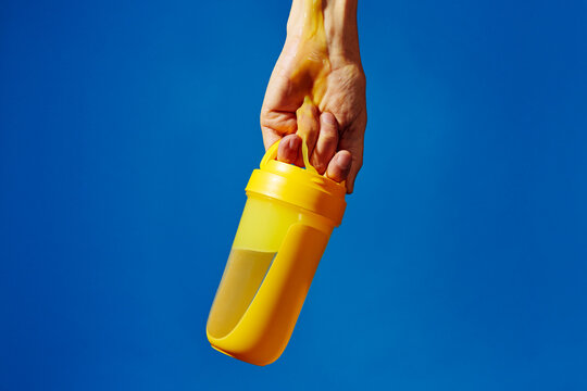 Hand holding yellow shaker bottle with protein spill against blue background