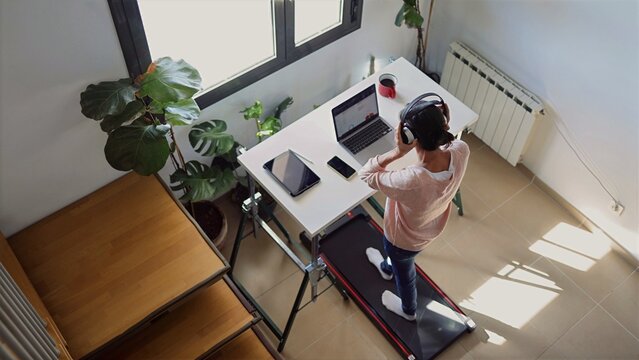 Woman working from home using a treadmill desk while walking, wearing headphones and looking at a laptop, staying active during remote work