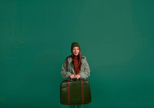 Smiling woman in fur coat with green suitcase against green background